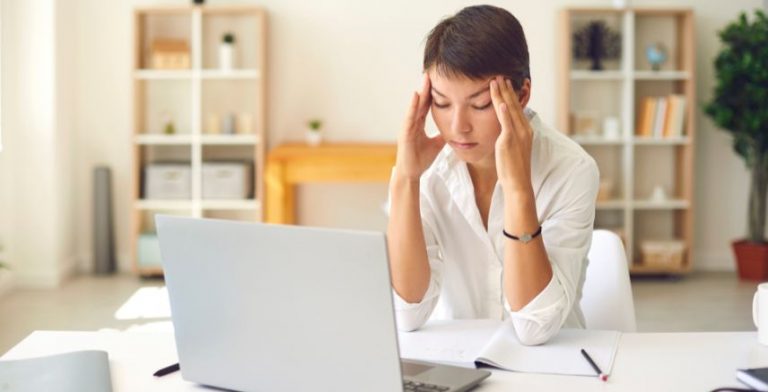 A woman sitting at a table in front of a laptop. She's holding her hands on both sides of her forehead and looks stressed.