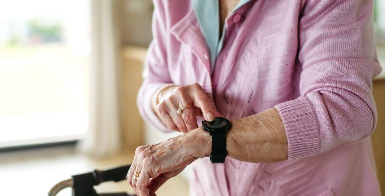 An elderly woman in a pink shirt touching her fall alert watch on her wrist.