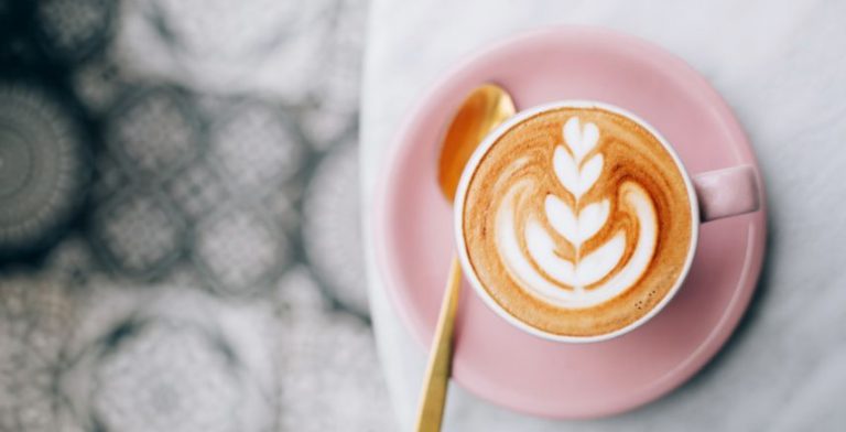 A latte with art in the foam, on a small pink plate with a gold spoon.